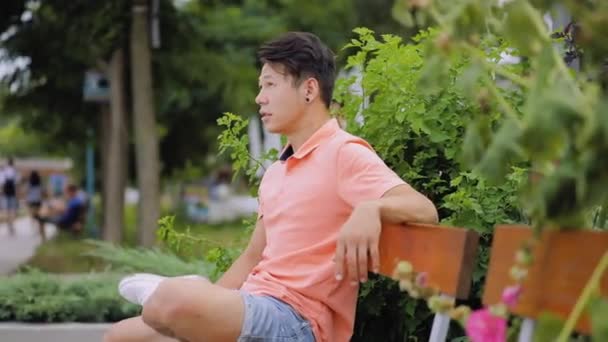 Азиатский мужчина с музыкой в голове сидит на городской скамейке и улыбается — стоковое видео