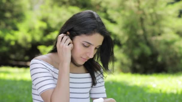 Молодая женщина вставила беспроводные воздушные стручки в уши, сидя на траве — стоковое видео