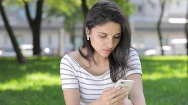 Genç kadınla çimenlerin üzerinde oturan smartphone kullanarak kablosuz airpods