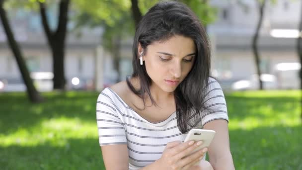 草の上に座ってのスマート フォンを使用して無線の airpods を持つ若い女性 — ストック動画