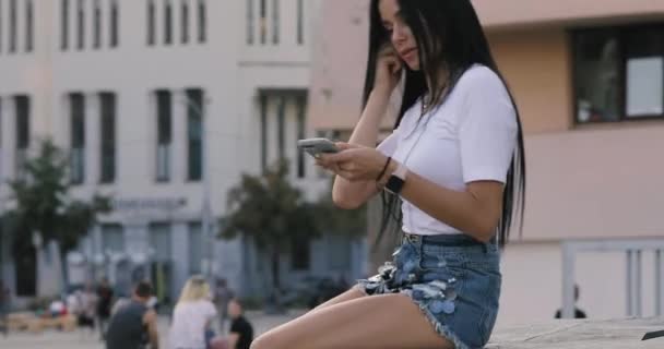 Mulher coloca fones de ouvido gostando de ouvir música sentada na praça da cidade — Vídeo de Stock