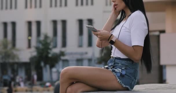 Mulher coloca fones de ouvido gostando de ouvir música sentada na praça da cidade — Vídeo de Stock