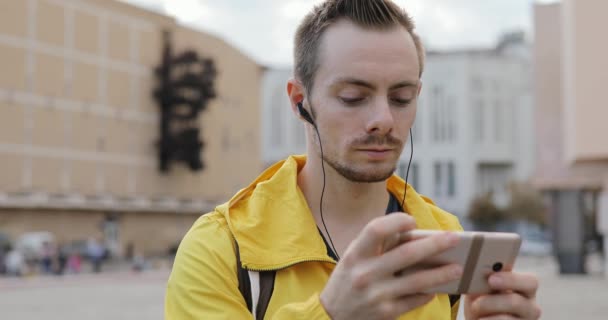 Jonge man in gele jas luisteren naar muziek in de oortelefoons op stedelijke stadsplein — Stockvideo