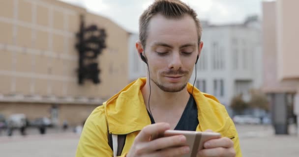 ชายหนุ่มในแจ็คเก็ตสีเหลืองสนุกกับการฟังเพลงในหูฟังในเมือง — วีดีโอสต็อก