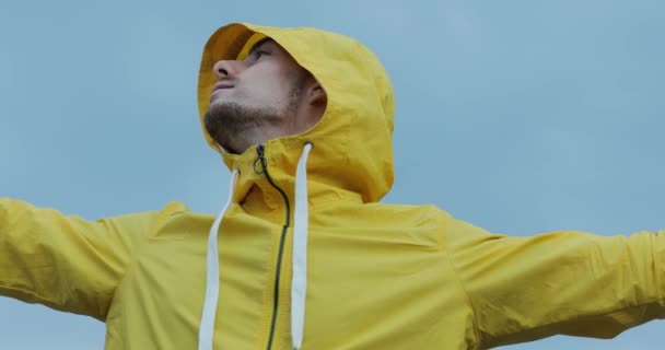 Hombre de chaqueta amarilla con las manos levantadas filmando a sí mismo por teléfono inteligente alrededor del cielo nublado — Vídeo de stock