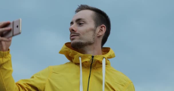 Człowiek w żółtą kurtkę z uniesionymi rękoma filmowanie siebie za pomocą smartfonów wokół pochmurnego nieba tło — Wideo stockowe