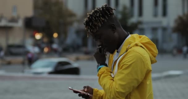 Hombre africano con chaqueta amarilla se pone auriculares en la calle de la ciudad escuchando música — Vídeos de Stock