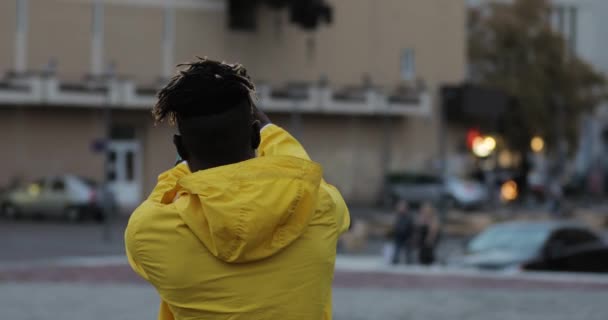 Hombre en rompevientos amarillo fotografiando plaza de la ciudad por la noche por teléfono celular, vista trasera — Vídeo de stock