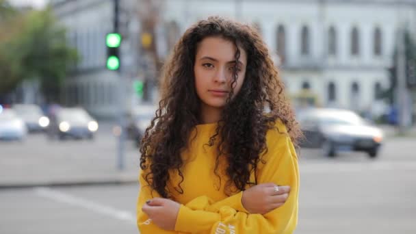 Glimlachend leuk krullend tiener meisje portret met arm stak in gele trui, stedelijke stad achtergrond — Stockvideo