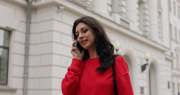 Happy woman in red blouse talking by smartphone going in city street — Stock Video