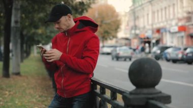 Çit, yolda arka plan üzerine eğilerek City akıllı telefon kullanarak kırmızı ceketli genç adam