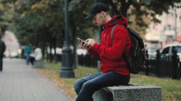 Junger Mann Roter Jacke Sitzt Mit Smartphone Auf Bank Gasse — Stockvideo