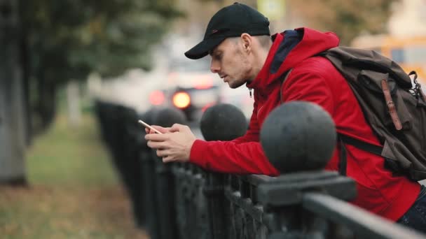 赤いジャケットの背景に道路フェンスに寄りかかって市でスマート フォンを使用しての若い男 — ストック動画