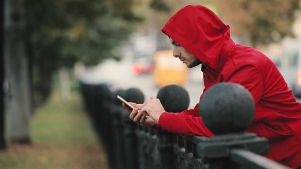 市では 背景に道路のフェンスに寄りかかってアプリ スマート フォンを使用して赤のジャケットの若い男 — ストック動画