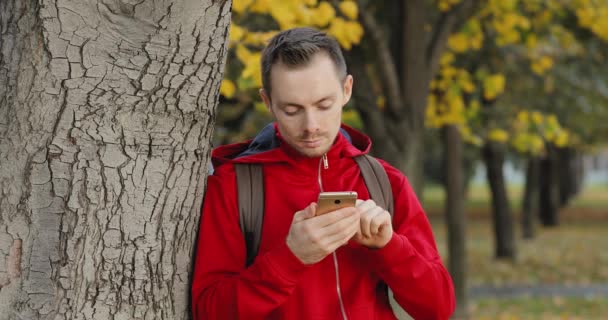 Mann i rød jakke med smarttelefon som lener seg mot trestammen om høsten – stockvideo