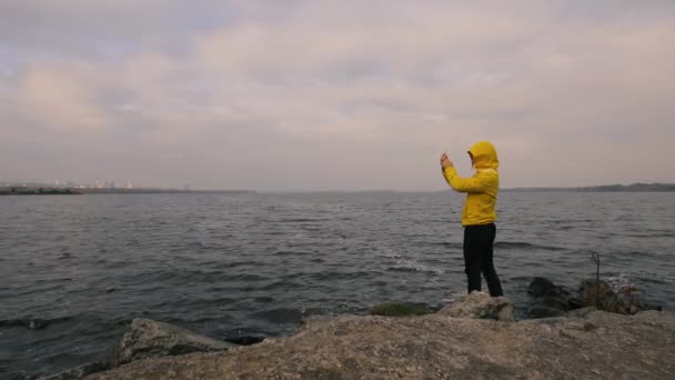 Путешественник в желтой куртке, гуляющий один на реке, сфотографируйся со смартфоном — стоковое видео