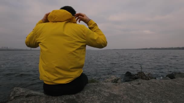 Solitudine, uomo in giacca gialla indossare cappuccio e pensare seduto al fiume — Video Stock
