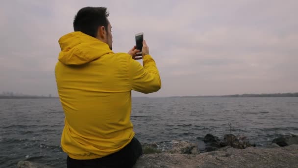 Tek başına nehir kıyısında oturmak sarı ceketli adamı gezgin hiking, panoramik bir video çekmek smartphone tarafından — Stok video