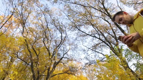 Человек в желтой куртке ходит со смартфоном в осеннем лесу — стоковое видео