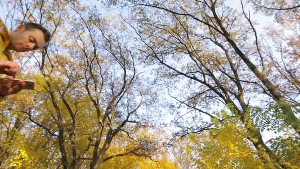 Hombre de chaqueta amarilla caminando con smartphone en el bosque otoñal — Vídeos de Stock