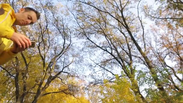 Hombre de chaqueta amarilla caminando con smartphone en el bosque otoñal — Vídeos de Stock