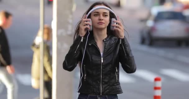 Modern woman puts on headphones while walking along city street, listening to music — Stock Video