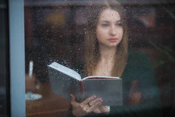 Nainen lukemassa kirjaa kahvilassa, näkymä märän ikkunan läpi sateisena päivänä — kuvapankkivalokuva