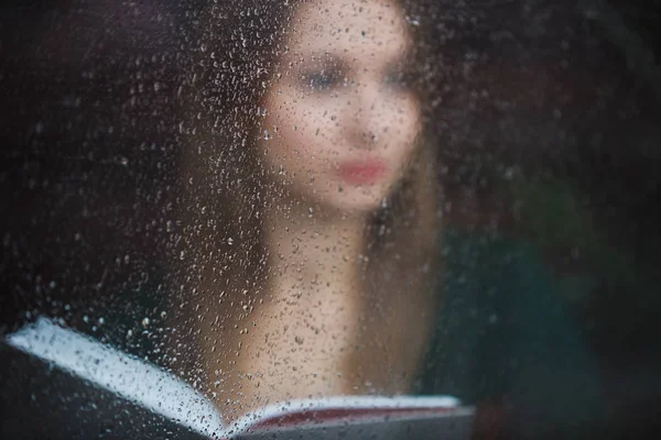 Mulher lendo livro no café, vista através da janela molhada no dia chuvoso — Fotografia de Stock