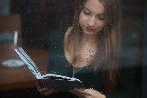 Kadın okuma kitabı kafede, ıslak camdan yağmurlu gün görüntülemek. — Stok fotoğraf