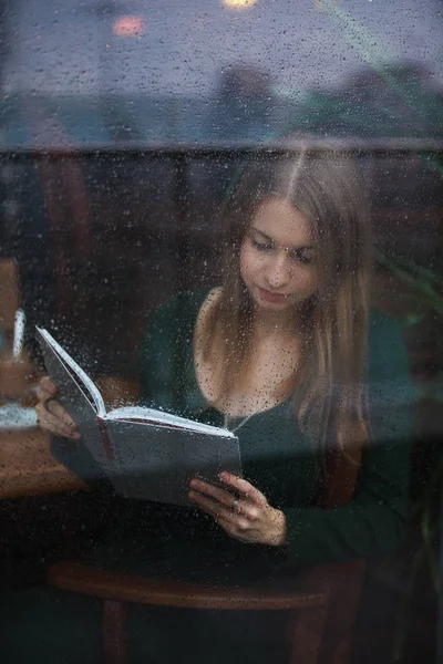 Kadın okuma kitabı kafede, ıslak camdan yağmurlu gün görüntülemek. — Stok fotoğraf
