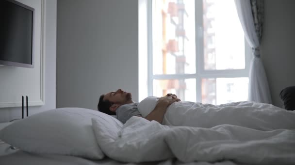 Homem acorda de manhã cedo em sua cama, esticando os braços — Vídeo de Stock