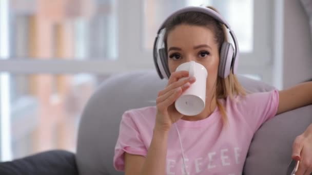 Mujer sonriente relajante en el sofá de casa con taza de té y buena música — Vídeos de Stock