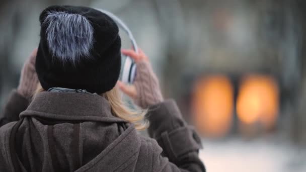 Вид ззаду на невпізнавану жінку, яка слухає музику в навушниках в зимовому вечірньому місті — стокове відео