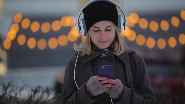 Зима, женщина думает и пишет сообщение на своем смартфоне в вечернем городе — стоковое видео