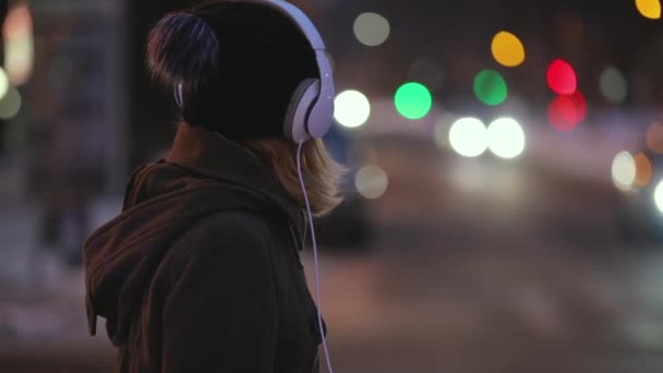 Mujer sola en auriculares listos para cruzar una calle, ciudad luces noche — Vídeo de stock