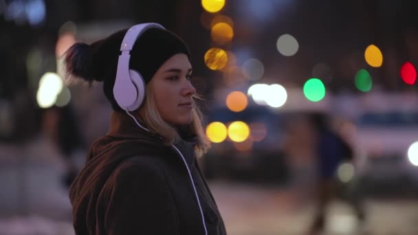 Mulher sozinha em fones de ouvido pronto para atravessar uma estrada, noite luzes da cidade — Vídeo de Stock