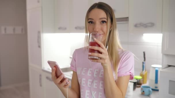 Feliz mañana, mujer bebiendo jugo de pie en la cocina escuchando música en casa — Vídeos de Stock