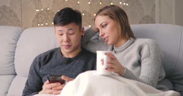 Mladý pár pomocí chytrého telefonu sedí na gauči doma — Stock video