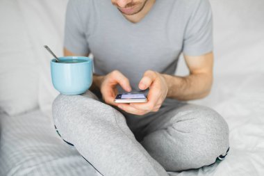 Sabah bir yatakta, cep telefonu, bacağını bardakta çay dengeleme kullanan adam