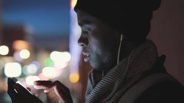 Primer plano del hombre africano uso de teléfono inteligente en los auriculares por la noche — Vídeos de Stock