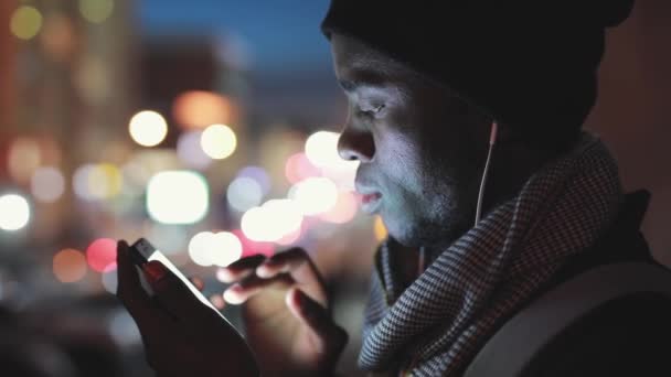 Primer plano del hombre africano uso de teléfono inteligente en los auriculares por la noche — Vídeo de stock