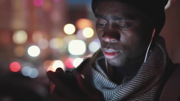Primer plano del hombre africano uso de teléfono inteligente en los auriculares por la noche — Vídeo de stock