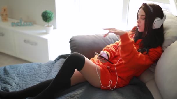 Милая девушка слушает музыку в наушниках на домашнем диване — стоковое видео