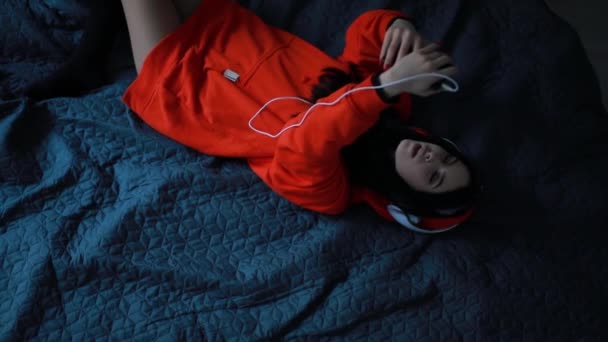 Девушка слушает музыку в наушниках, лежащих на кровати, вид сверху — стоковое видео