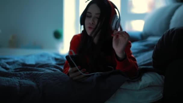 Девушка слушает музыку в наушниках, лежащих на кровати — стоковое видео
