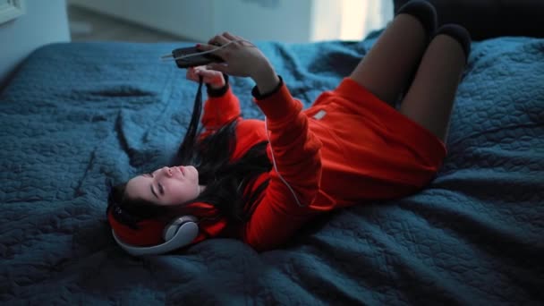 Chica escuchando música en los auriculares acostados en la cama — Vídeos de Stock