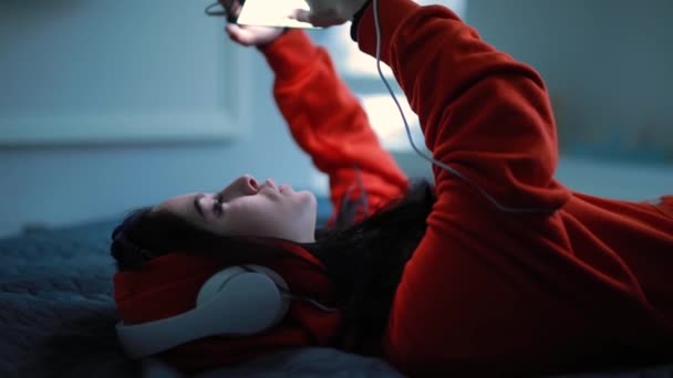 Flicka lyssnar musik i hörlurar liggande på sängen — Stockvideo