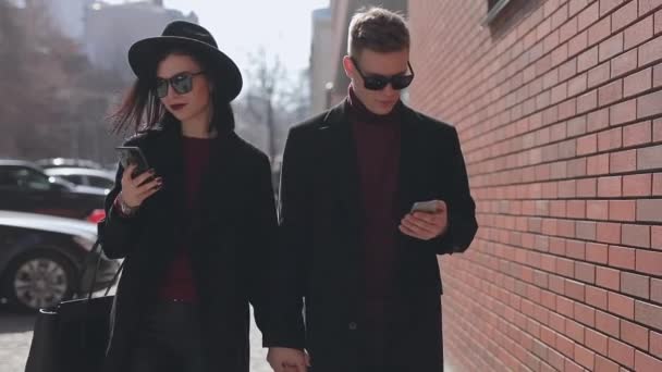 Stylish couple looking down into their smartphones walking along city street — Stock Video