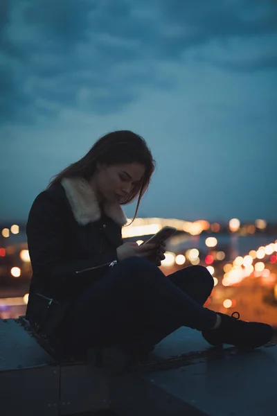 Siluetti yksin nainen istuu älypuhelin katolla, kaupunki taustalla — kuvapankkivalokuva