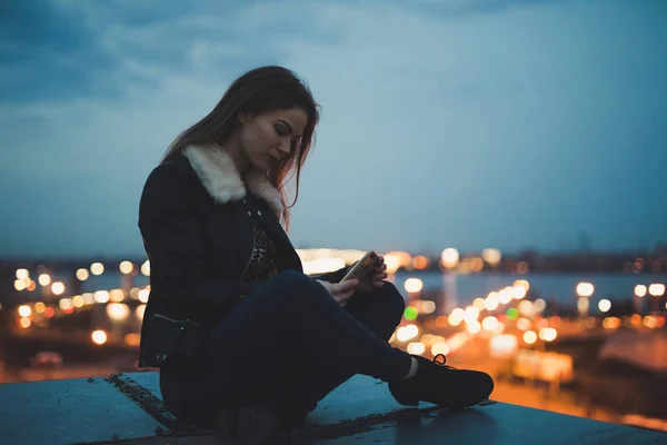 Silueta de mujer sola sentada con teléfono inteligente en el techo, ciudad en el fondo —  Fotos de Stock